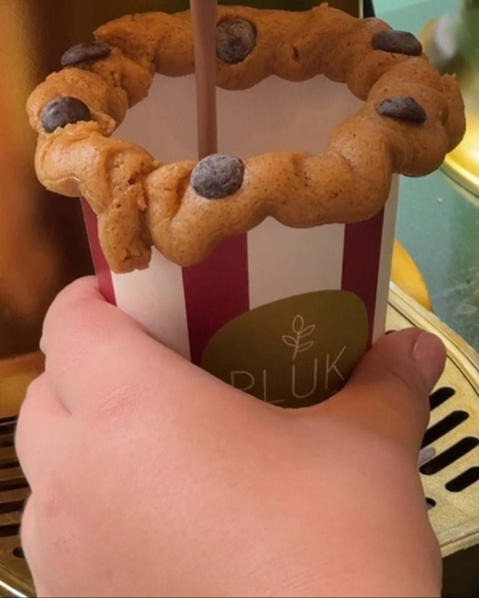 Hot chocolate pouring from a dispenser into a red and white striped paper cup with a ring of chocolate chip cookie dough around the rim.