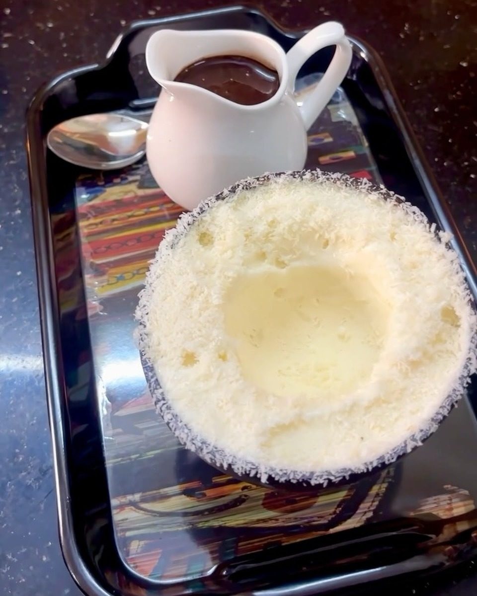 Ice cream in a coconut shell cup on a black tray, with a white jug of thick hot chocolate and a spoon beside it.