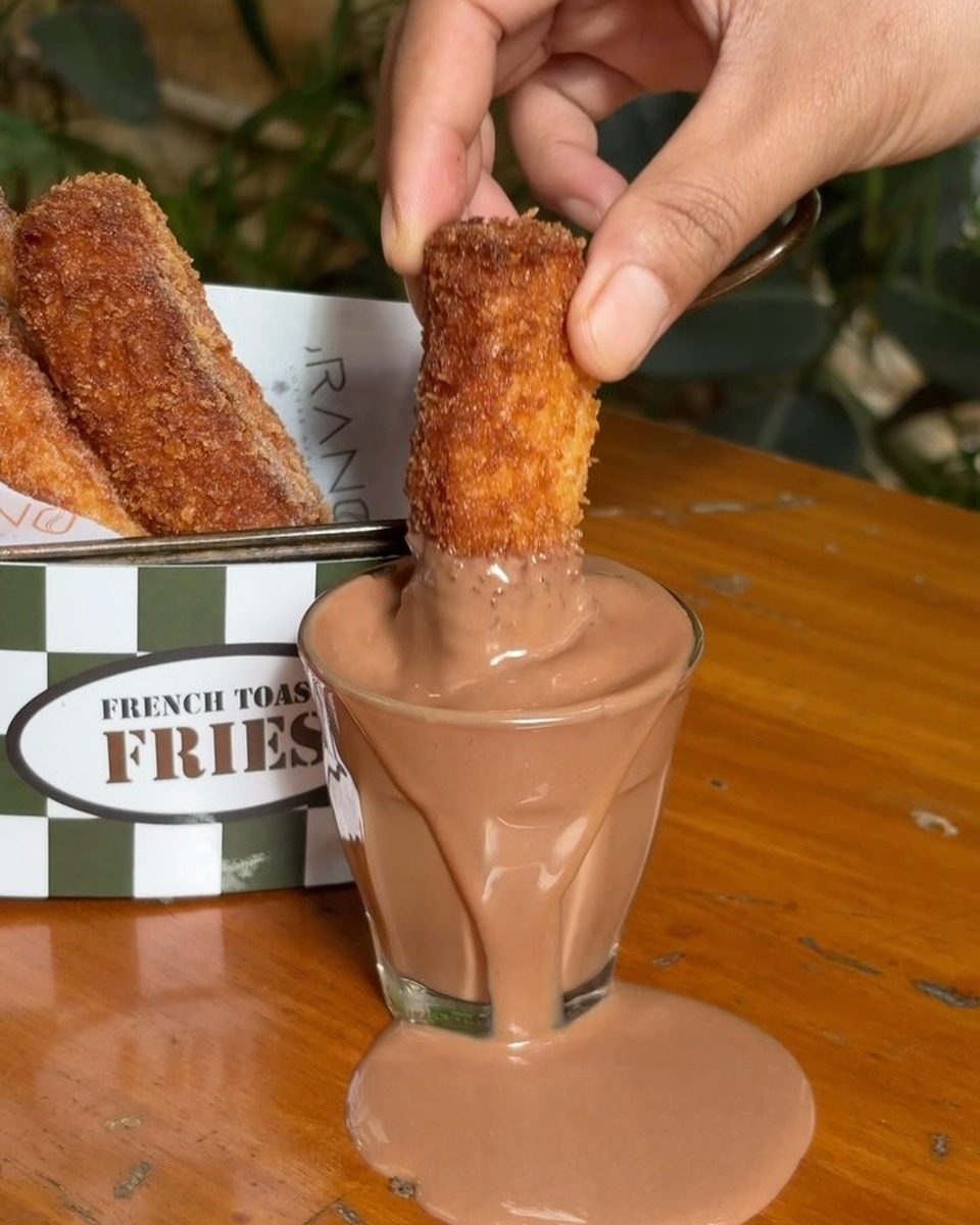 A french toast fry dipped into a small glass of chocolate that runs over the rim, with more fries in a paper box behind it.