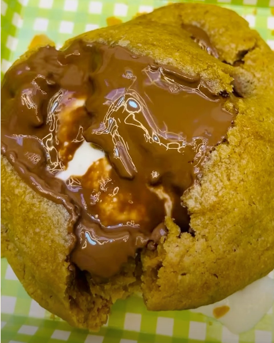A thick baked cookie broken open on a green and white checked paper tray. Melted milk chocolate and a white marshmallow run out of the centre of the cookie.