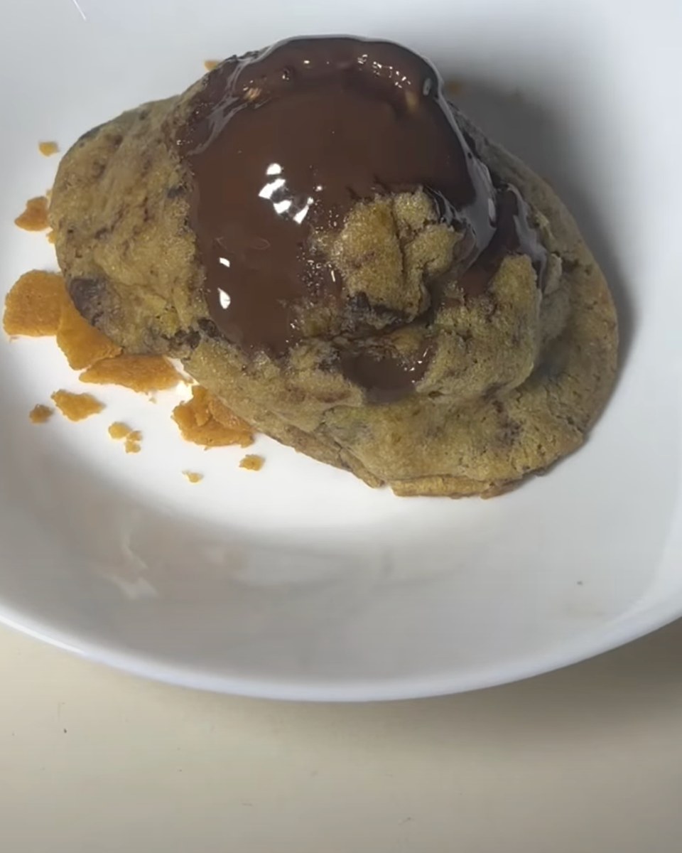 A thick chocolate chip cookie in a white bowl with melted dark chocolate poured over the top and a few crumbs of crunch around it.