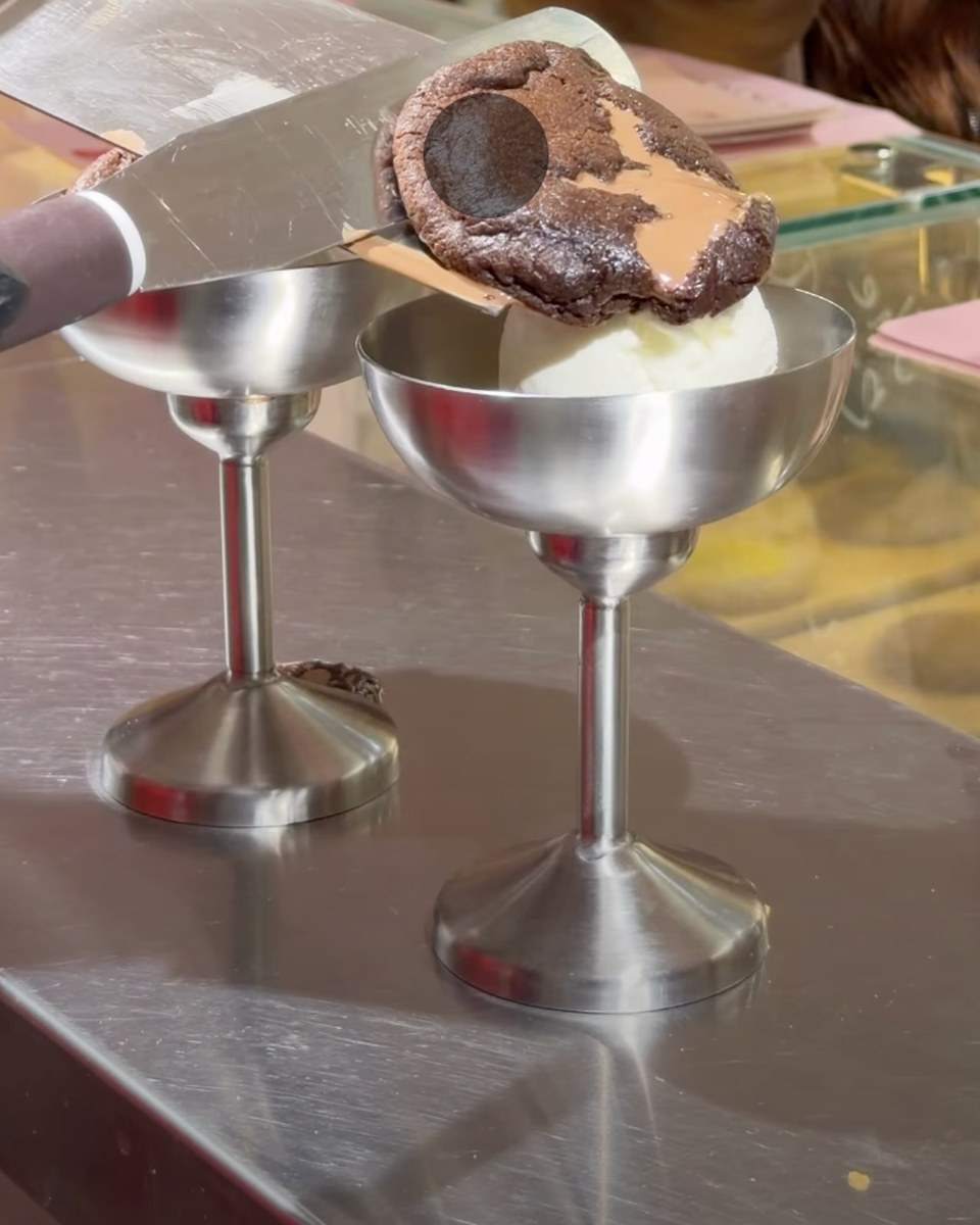 A baked molten cookie with a tan, biscoff-coloured spread across the top, lifted on a metal spatula over a scoop of vanilla ice cream in a steel dessert cup. A second steel cup stands on the steel counter beside it.
