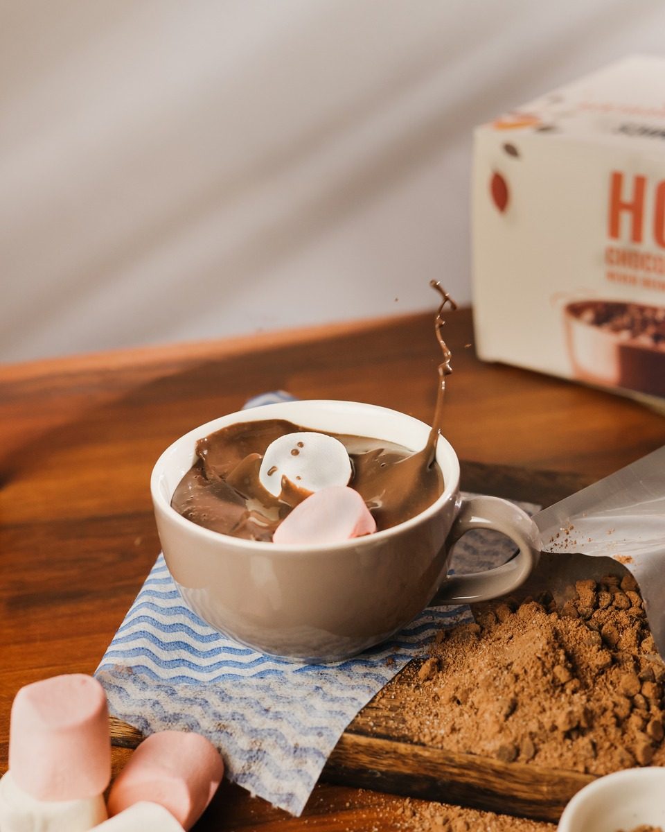 A cup of thick hot chocolate with marshmallows on a wooden board, with cocoa and a hot chocolate box behind it.