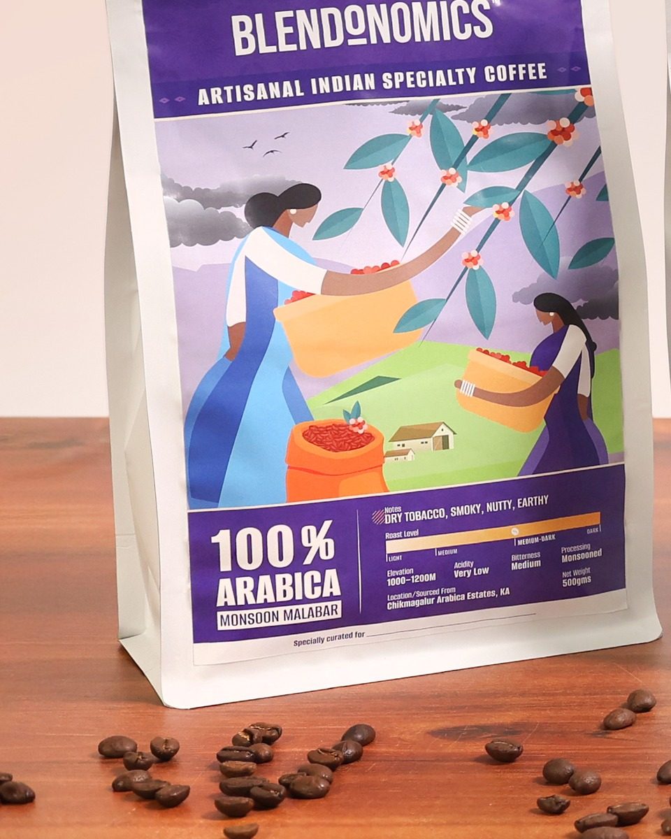 A Blendonomics Monsoon Malabar coffee pack, 100 percent Arabica, on a wooden table with roasted beans.
